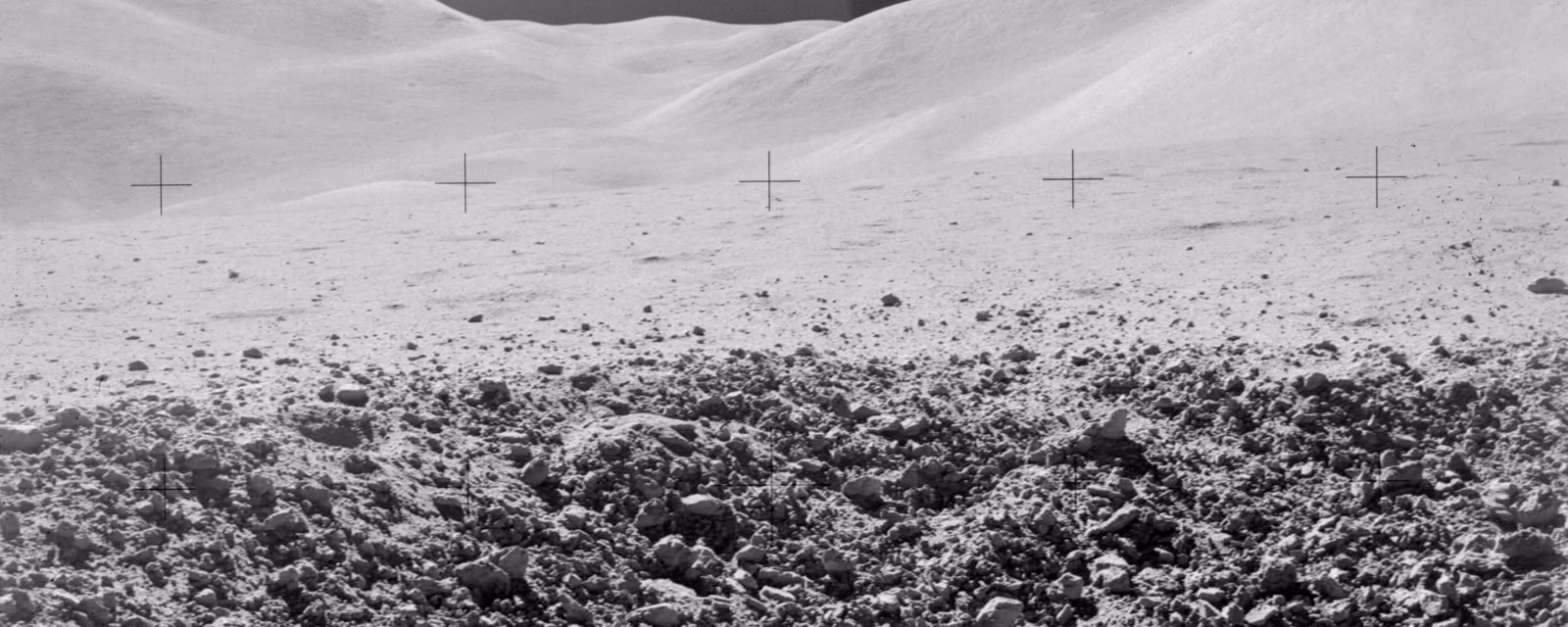 A black-and-white Apollo 15 photograph of the Moon's surface: a field of loose rock in the foreground, with pale rounded hills along the horizon under a dark sky.